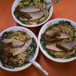 朱華楼 ・東深津店　〒721-0974 広島県福山市東深津町１丁目９−６３　しゅうかろう　ラーメン らーめん いなり　愛知 名古屋 Alto e Diritto altoediritto アルトエデリット 国道２号線と１８２号線の交わる交差点の西に福山通運のある交差点を北上。高架を通り抜け２００メートル程北上した左側（ゴルフショップダイナマイト駐車場の道路を挟んで向かい側）東福山駅から1,362m　中華そば 専門店 広島県 ひろしま 尾道ラーメン 尾道ラーメン。広島県東部の尾道市から福山市で広がるご当地ラーメン。鶏ガラ中心の醤油スープに、大粒の背脂が浮かぶのが特徴。瀬戸内産のいりこを使っているお店も多い。名前は全国的に知られているが、実は現地には20～30軒しかない珍しいご当地ラーメンだ。一番の有名店は尾道市の「朱華園」。1947年に屋台から始まったお店で、全国からお客の集まる行列店だったが、2019年6月に惜しまれつつも閉店。ラーメン業界的には大きなニュースになった。「朱華園」は閉店してしまったものの、そのDNAを受け継ぐお店がいくつかある。今回ご紹介するのは、そんな「朱華園」の流れを汲む「朱華楼」。福山市内で3店舗を展開しているが、東深津店が代表として語られることが多い。メニューは「中華そば」と「いなり寿司」のみ。中華そばは600円だ。